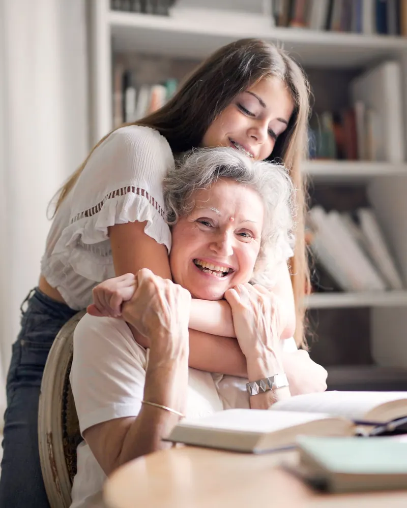 Pflegekraft und Seniorin sitzen nebeneinander und lachen gemeinsam