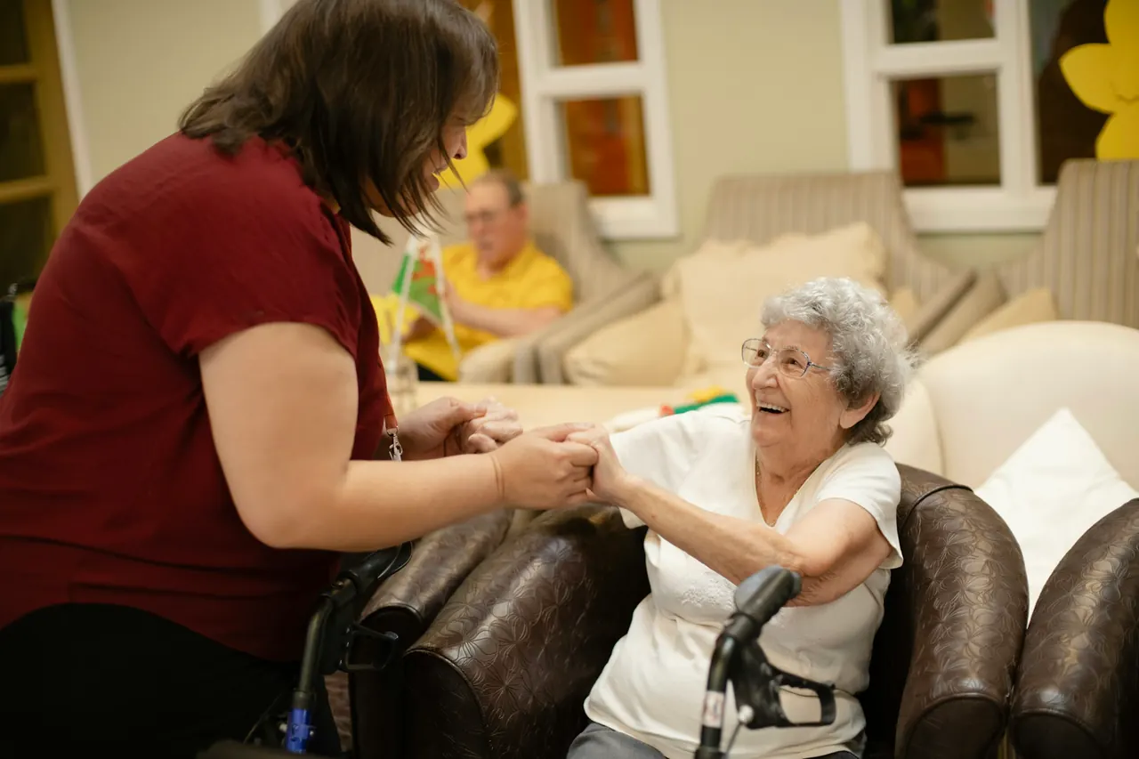 Pflegekraft hält die Hand einer Seniorin in ihrem Wohnzimmer – ein ruhiges Gespräch bei warmem Licht