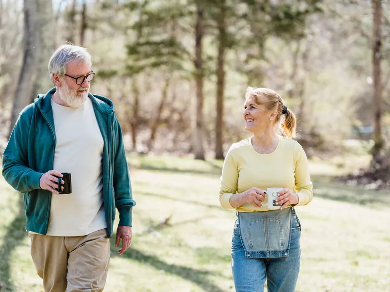 Älterer Mann und eine Frau gehen mit Bechern in der Hand durch einen sonnigen Wald