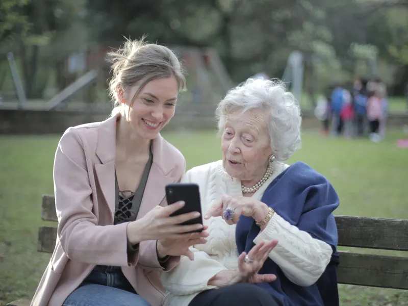 Junge Frau und Seniorin schauen lachend gemeinsam auf ein Smartphone auf einer Parkbank